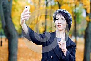 Beautiful woman taking selfie by smartphone and having fun in autumn city park, fall season, yellow leaves