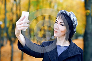 Beautiful woman taking selfie by smartphone and having fun in autumn city park, fall season, yellow leaves