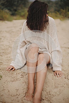Beautiful woman with tanned legs in sand sitting on sandy beach with green grass, carefree tranquil moment. Stylish young sexy