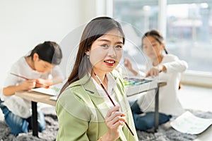 beautiful woman teacher smile, happy, look at camera, hold color paintbrush in education classroom. asian beautiful children happy
