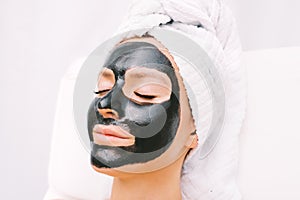 Beautiful woman with towel on her head relaxing with black charcoal facial mask