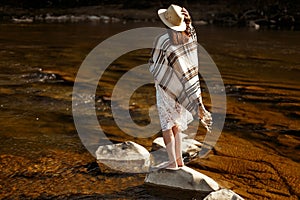 beautiful woman traveler back standing on rocks in river, wearing hat and poncho, boho travel concept