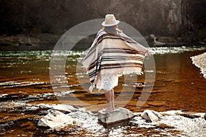 Beautiful woman traveler back standing on rocks in river, wearing hat and poncho, boho travel concept