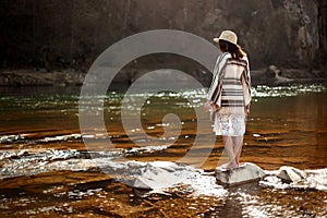 beautiful woman traveler back standing on rocks in river, wearing hat and poncho, boho travel concept