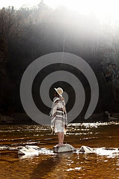 beautiful woman traveler back standing on rocks in river, wearing hat and poncho, boho travel concept, amazing view