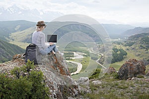 Beautiful woman traveler in a hat in mountains with a laptop.