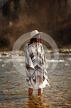 beautiful woman traveler wearing hat and poncho standing in water of river, stylish outfit, boho travel concept