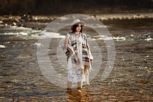 beautiful woman traveler wearing hat and poncho standing in water of river, stylish outfit, boho travel concept