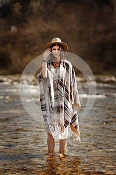 beautiful woman traveler wearing hat and poncho standing in water of river, stylish outfit, boho travel concept