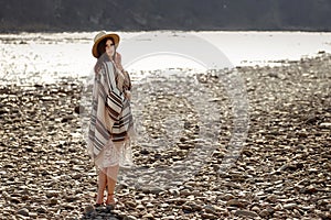 Beautiful woman traveler wearing hat and poncho walking at river beach, american outfit, boho concept, space for text