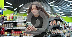 Beautiful woman use smartphone in supermarket. Woman buyer using mobile phone application standing with shopping trolley