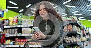 Beautiful woman use smartphone in supermarket. Woman buyer using mobile phone application standing with shopping trolley