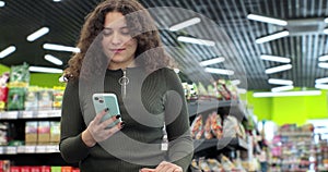 Beautiful woman use smartphone in supermarket. Woman buyer using mobile phone application standing with shopping trolley