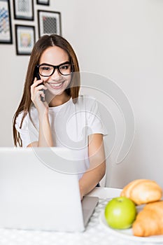 Beautiful woman is using a laptop, talking on the mobile phone and smiling while cooking in kitchen at home