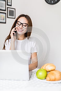 Beautiful woman is using a laptop, talking on the mobile phone and smiling while cooking in kitchen at home