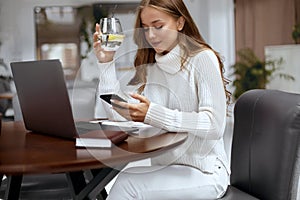 Beautiful woman using smartphone while working with laptop and holding glass of water.