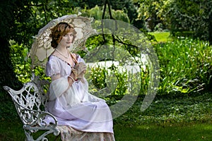 Beautiful woman in vintage dress sitting on ornate metal bench holding parasol next to river photo