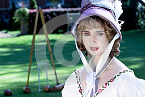 Beautiful woman in vintage Georgian dress and bonnet sitting on lawn in front of stately home.