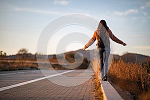 Beautiful woman walking and balancing on street curb or curbstone during sunset