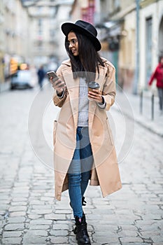 Beautiful smiling woman walking on city street from work with coffee cup and texting on mobile phone