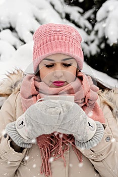 Beautiful woman in warm winter clothes holding cup drinking hot tea or coffee outdoors in snowy day
