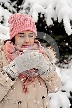 Beautiful woman in warm winter clothes holding cup drinking hot tea or coffee outdoors in snowy day