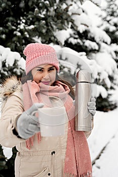 Beautiful woman in warm winter clothes holding thermos and drinking hot tea or coffee outdoors in snowy day