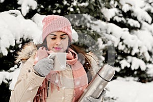 Beautiful woman in warm winter clothes holding thermos and drinking hot tea or coffee outdoors in snowy day