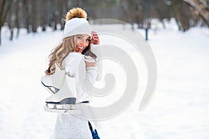 Beautiful woman weared in white sweater and hat with ice skates on the back walks in winter snowy park.
