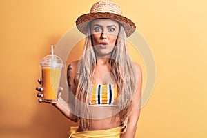 Beautiful woman wearing bikini and hat drinking healthy orange juice over yellow background scared and amazed with open mouth for