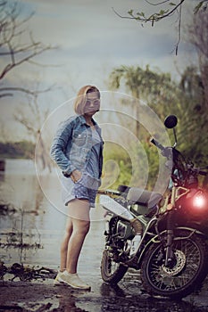 beautiful woman wearing blue jeans jacket standing beside enduro motorcycle against colorful natural background