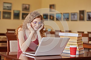 Beautiful woman wearing burgundy casual t shirt and glasses reading text via laptop or searching necessary information, using