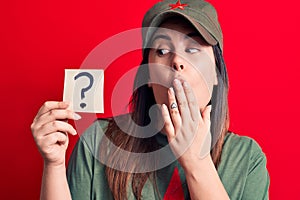 Beautiful woman wearing cap with red star communist symbol holding question mark reminder covering mouth with hand, shocked and