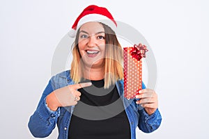 Beautiful woman wearing Christmas Santa hat holding gift over isolated white background very happy pointing with hand and finger