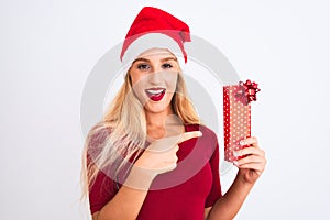 Beautiful woman wearing Christmas Santa hat holding gift over isolated white background very happy pointing with hand and finger