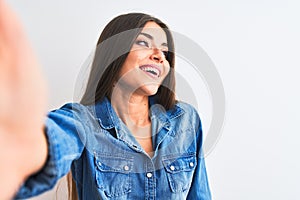 Beautiful woman wearing denim shirt make selfie by camera over isolated white background looking away to side with smile on face,