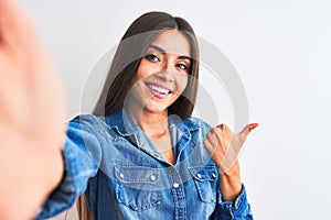 Beautiful woman wearing denim shirt make selfie by camera over isolated white background Pointing to the back behind with hand and