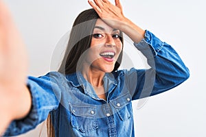 Beautiful woman wearing denim shirt make selfie by camera over isolated white background surprised with hand on head for mistake,