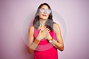 Beautiful woman wearing funny pink heart shaped sunglasses over pink isolated background smiling with hands on chest with closed