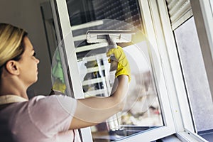 Beautiful Woman wearing Protective Gloves Cleaning Window by spraying Cleaning Products and wiping with Sponge