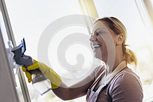 Beautiful Woman wearing Protective Gloves Cleaning Window by spraying Cleaning Products and wiping with Sponge