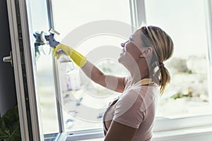 Beautiful Woman wearing Protective Gloves Cleaning Window by spraying Cleaning Products and wiping with Sponge