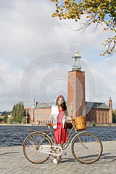 Beautiful woman wearing red dress dress holding a vintage bicycle in front of Stockholm City Hall