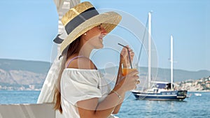 Beautiful woman wearing straw hat, sits in a gazebo on the wooden sea pier. She enjoys her orange juice or cocktail