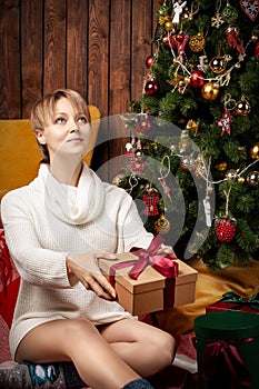 Beautiful woman wearing a warm sweater and socks, sitting near tha Christmas tree holding gift boxes