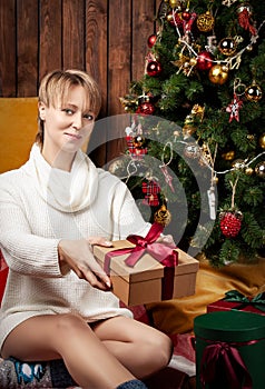 Beautiful woman wearing a warm sweater and socks, sitting near tha Christmas tree holding gift boxes