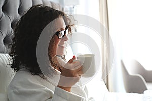 Beautiful woman in a white bathrobe with a cup of coffee on the bed at the hotel