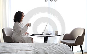 Beautiful woman in a white bathrobe with a cup of coffee on the bed at the hotel.