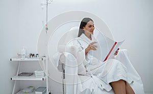 Beautiful woman in white bathrobe reading magazine while sitting in luxury wellness spa centre