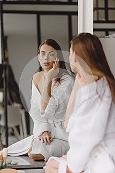 Beautiful woman in white bathrobe sitting in front of mirror in bathroom and putting undereye patches on face. photo